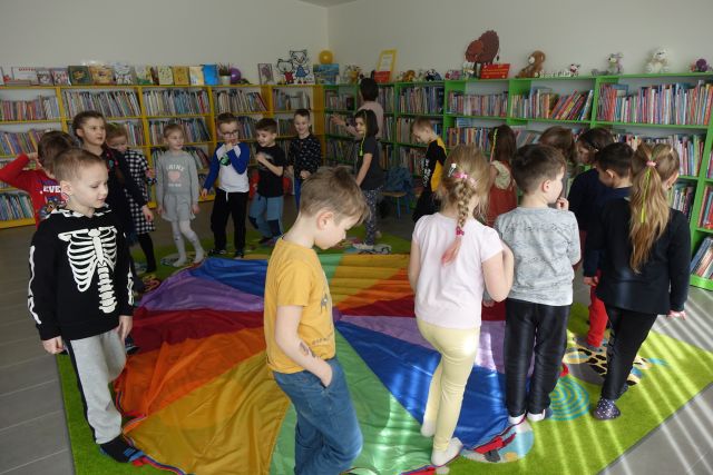 dzieci chodzą wokół kolorowej chusty, w tle regały z książkami