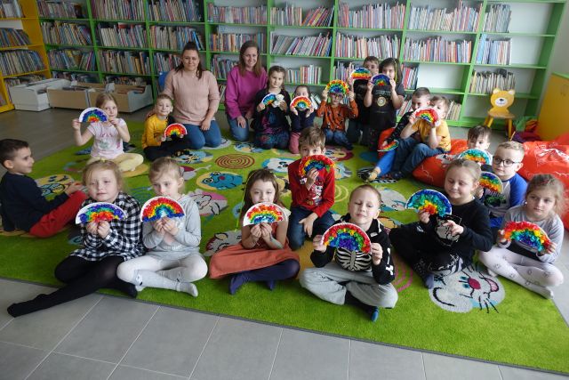 dzieci siedzą na zielonym dywanie i pozują do zdjęcia z wykonanymi z papierowych talerzyków i bibuły tęczami, w tle regały z książkami