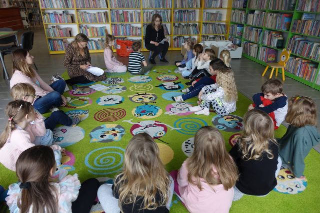 dzieci i dwie panie siedzą na zielonym dywanie w okręgu, przed nimi siedzi na krzesełku i czyta dzieciom, w tle regały z książkami