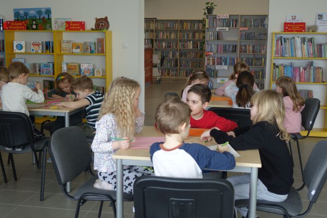 dzieci siedzą przy stolikach i rysują na kartkach, w tle kolorowe regały