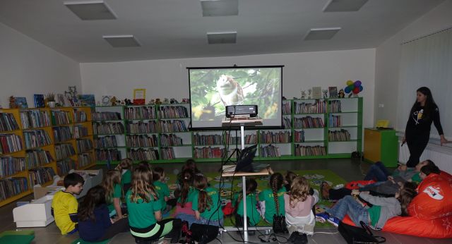 kobieta ubrana na czarno stoi przed dziećmi siedzącymi na dywanie i opowiada o sowach, z jej lewej strony ekran z wyświetlaną prezentacją