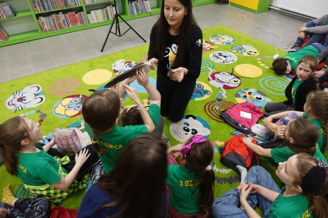 kobieta na czarno kuca na zielonym dywanie przed dziećmi i pokazuje im pióro sowy