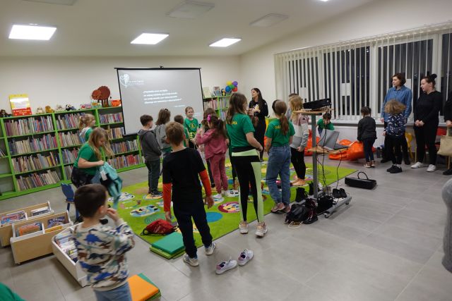dzieci i kobieta stoją na zielonym dywanie, przed nimi ekran