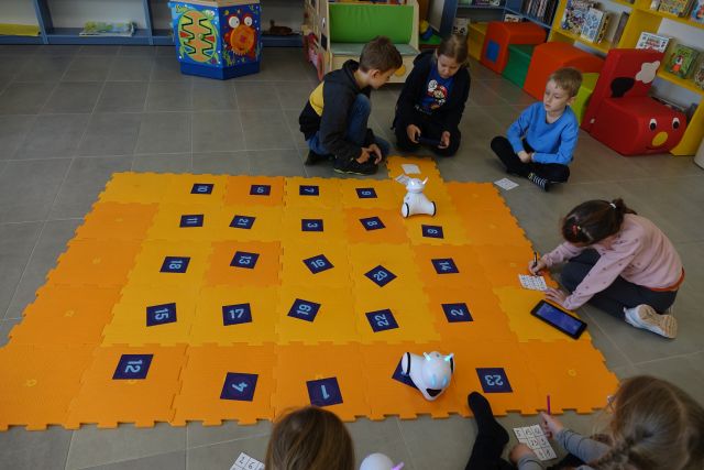 dzieci klęczą wokół pomarańczowych puzli, na których stoją małe białe roboty photon i kartki z liczbami