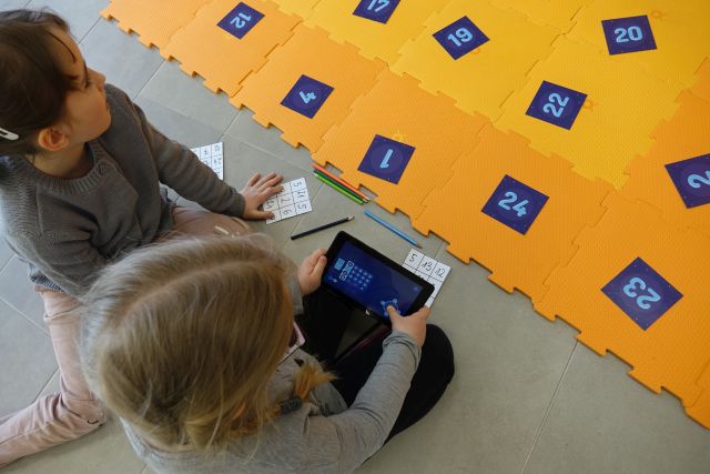 dwójka dzieci w tabletach przegląda aplikację, przed nimi pomarańczowa mata z puzli i na niej kartki z liczbami