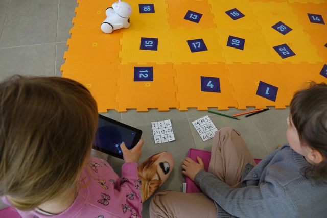 dwójka dzieci w tabletach przegląda aplikację, przed nimi pomarańczowa mata z puzli i na niej kartki z liczbami