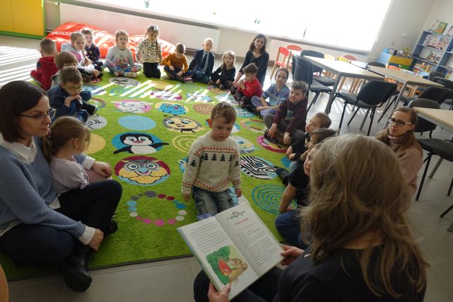 dzieci siedzą na zielonym dywanie i słuchają czytanej przez kobietę ubraną na czarno książkę, w tle okna