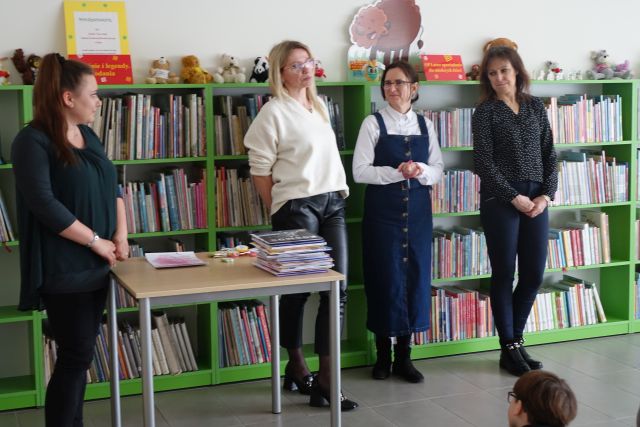 na tle regałów z książkami stoją 4 kobiety, pomiędzy pierwszą a drugą stoi stolik, na nim książki i dyplomy dla laureatów konkursu