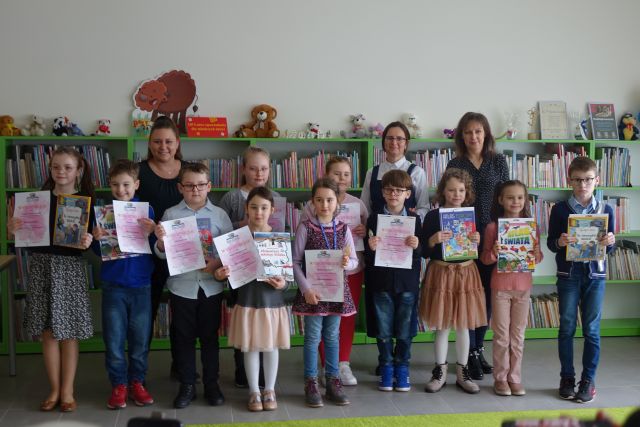 11 dzieci nagrodzonych w konkursie stoi z nagrodami książkowymi i dyplomami i pozuje do zdjęcia z nimi stoją 3 kobiety na tle regałów z książkami