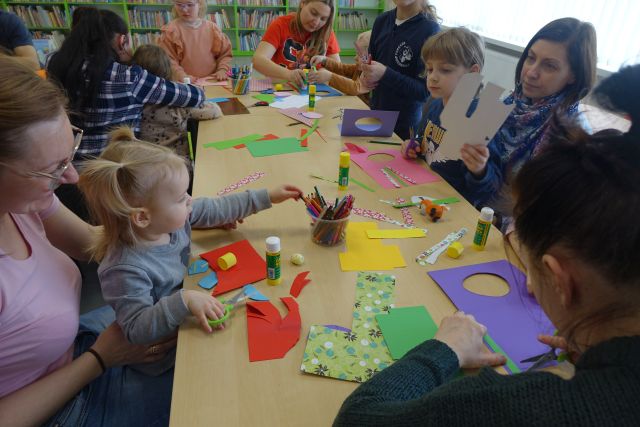 około 25 dzieci i dorosłych siedzi przy stołąch i robi kartki wielkanocne