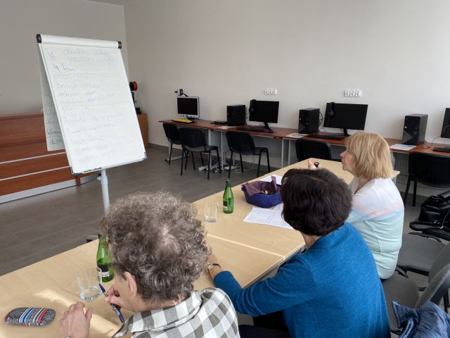 trzy kobiety siedzą przy stolikach i piszą na kartkach, przed nimi flipchart, w tle komputery na biurkach