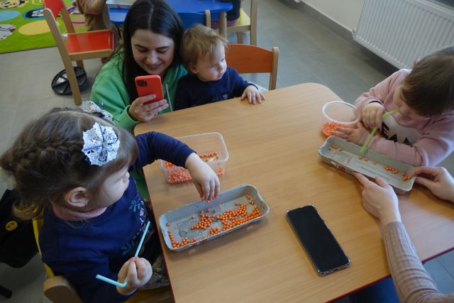 dzieci siedzą przy stołach i mieszają w koralikach, przy nich kuca kobieta z telefonem