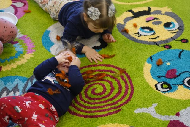 dzieci siedzą  i leżą na zielonym dywanie i bawią się piórkami