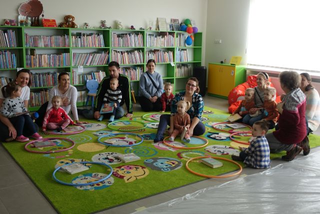 dzieci i rodzice siedzą na zielonym dywanie, pomiędzy nimi hula hop, w tle regały z książkami