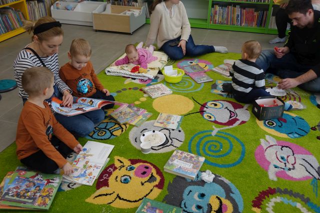 dzieci i rodzice siedzą na zielonym dywanie i przeglądają książki