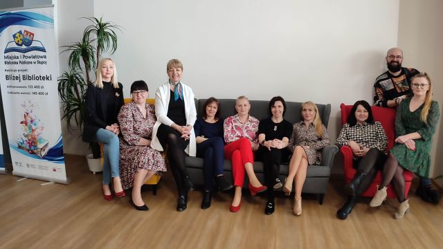 9 kobiet i mężczyzna siedzą na żółtym i czerwonym fotelu oraz siwej kanapie, z lewej strony baner z informacją o projekcie i logo biblioteki