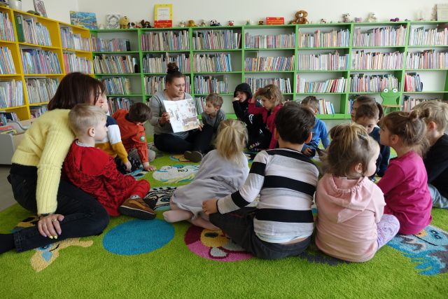 dzieci siedzą na kolorowym dywanie i patrzą na książkę pokazywaną przez bibliotekarkę
