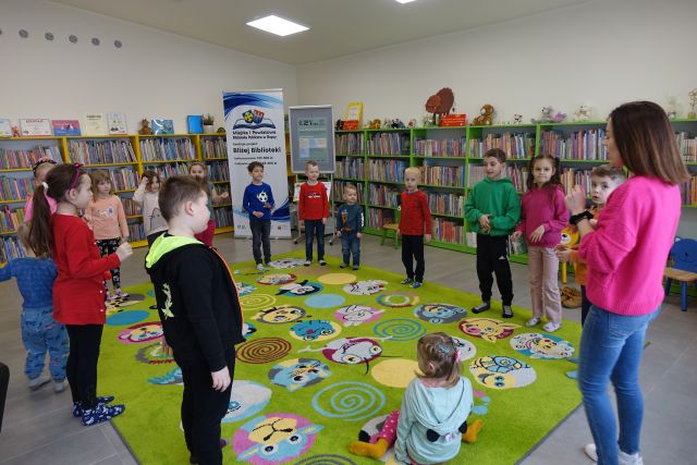 dzieci i kobieta stoją na zielonym dywanie, w tle regały z książkami