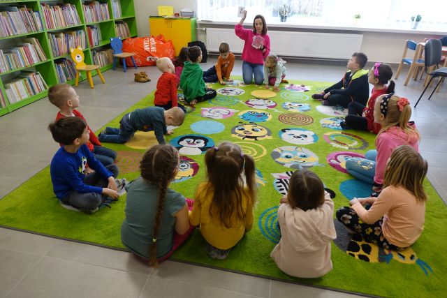 dzieci siedzą na dywanie, przed nimi siedzi kobieta w różowej bluzce i pokazuje kartkę