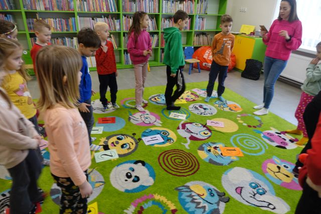 dzieci stoją na zielonym dywanie pomiędzy nimi kartki z cyframi, w tle regały z książkami