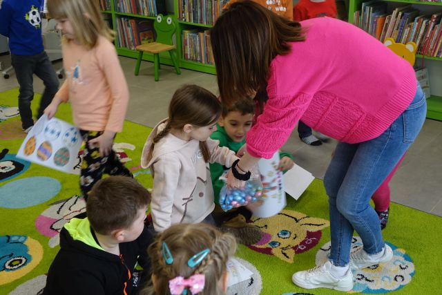 kobieta w różowej bluzce częstuje dzieci siedzące na dywanie słodyczami