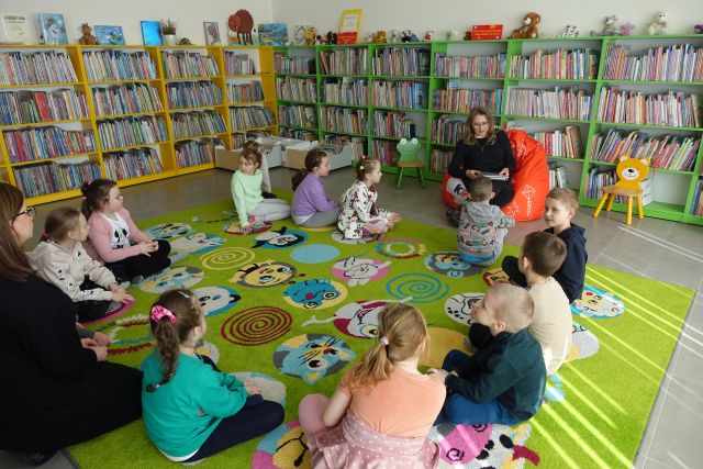13 dzieci siedzi w okręgu na zielonym dywanie i słucha jak kobieta siedząca na pomarańczowej pufie czyta, w tle regały z książkami