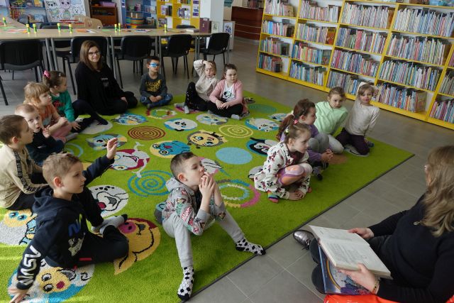 13 dzieci siedzi w okręgu na zielonym dywanie i słucha jak kobieta siedząca na pomarańczowej pufie czyta, w tle regały z książkami