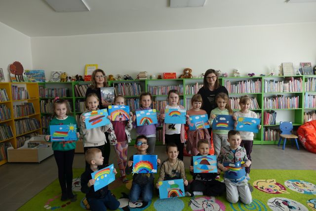 13 dzieci i dwie osoby dorosłe pozują do zdjęcia z wykonanymi przez siebie iwyklejonymi tęczami, w tle regały z książkami
