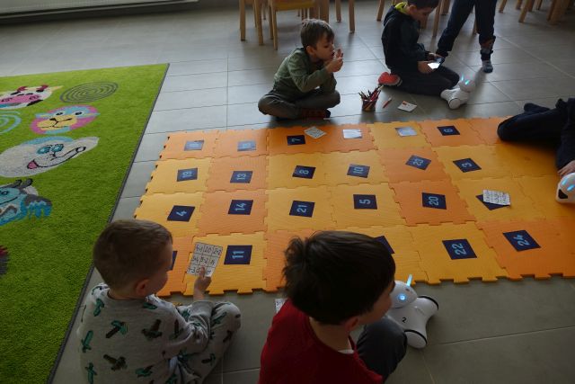 dzieci siedzą wokół pomarańczowej maty z puzli, na niej kartki z liczbami, obok roboty photon