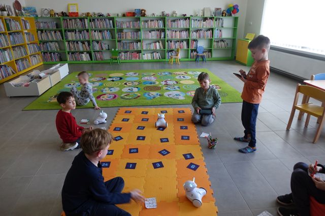 dzieci siedzą wokół pomarańczowej maty z puzli, na niej kartki z liczbami i roboty photon