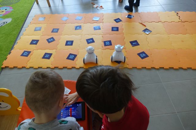 dzieci siedzą wokół pomarańczowej maty z puzli, na niej kartki z liczbami i roboty photon