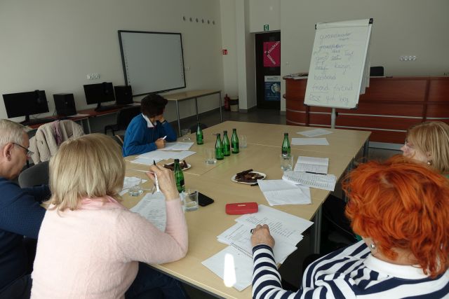 4 kobiety i 1 mężczyzna uczestniczą w zajęciach z języka angielskiego, na stołach kartki, długopisy, ptasie mleczko, woda, w tle sala komputerowa