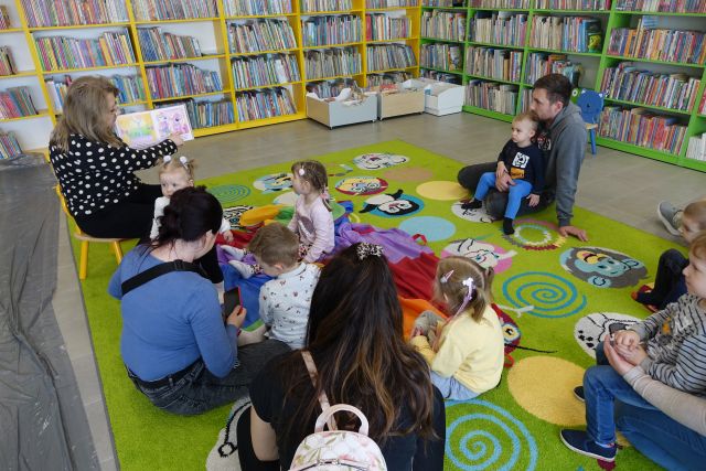 dzieci i rodzice siedzą na kolorowym dywanie i słuchają czytaną książkę przez biblkiotekarkę, w tle regały z książkami