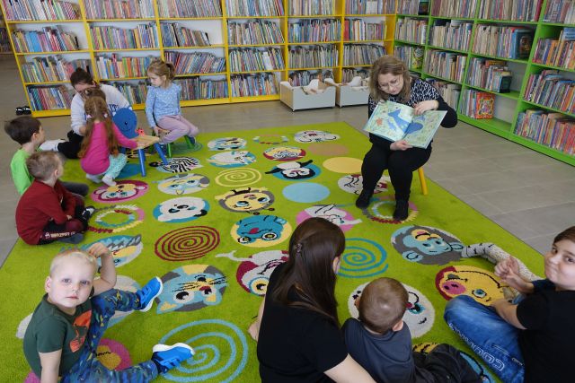 dzieci i rodzice siedzą na zielonym dywanie, przed nimi na małym krzesełku siedzi kobieta pokazująca książkę o delfinach, w tle regały z książkami