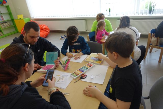 dzieci z rodzicami siedzą przy stolikach i malują delfiny