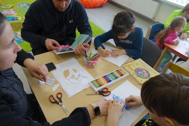 dzieci i rodzice siedzą przy stolikach i maluja delfiny