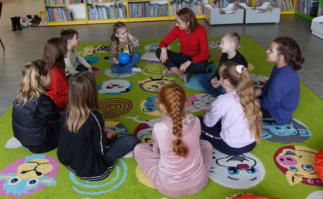 9 dzieci i kobietą siedzą w kole na zielonym dywanie, w tle regały z książkami