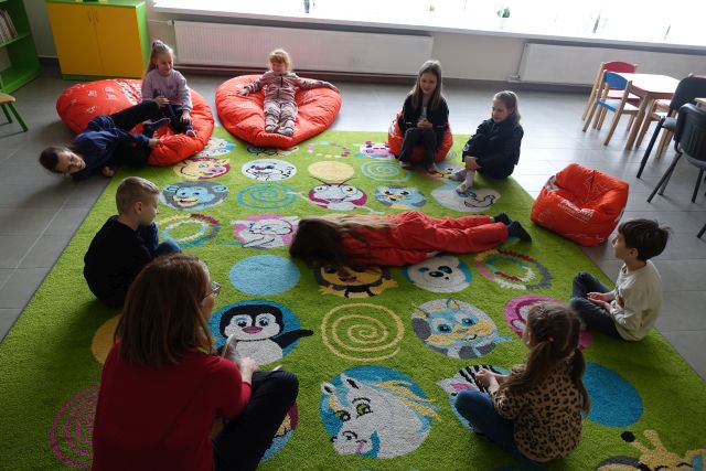 8 dzieci i kobieta siedzą na zielonym dywanie, jedno dziecko leży na brzuchu na środku dywanu