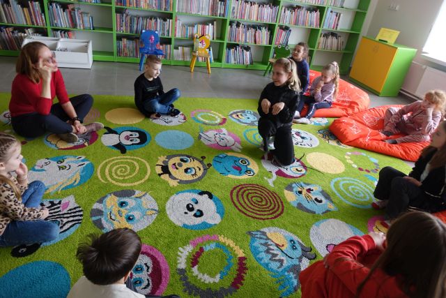 8 dzieci i kobieta siedzą na kolorowym dywanie, dziewczynka w środku kuca udając królika, w tle regały z książkami