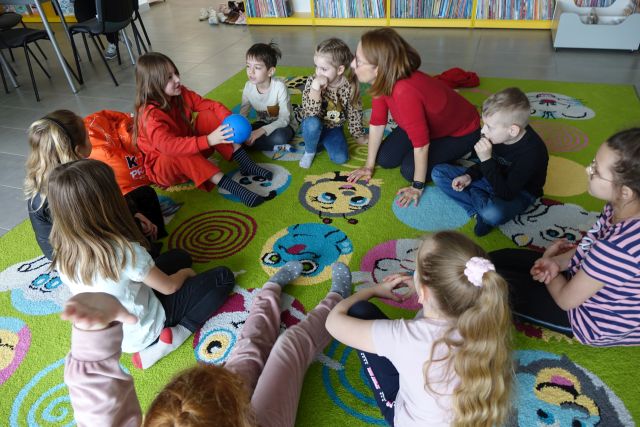 9 dzieci i kobietą siedzą w kole na zielonym dywanie, w tle regały z książkami