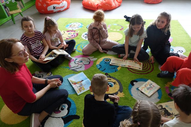 9 dzieci i kobietą siedzą w kole na zielonym dywanie, w tle regały z książkami, pomiędzy dziećmi leżą na dywanie książki