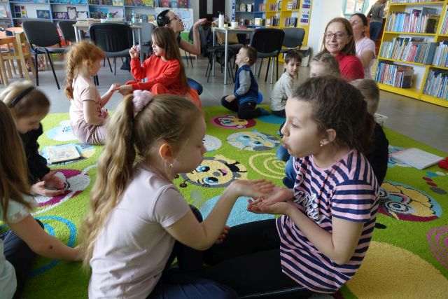 dzieci i 2 kobiety siedzą na dywanie i dmuchają piórka