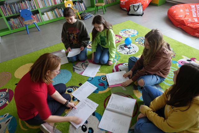 4 dziewczyny i kobieta siedzą na kolorowym dywanie, przed nimi leżą zapisane kartki