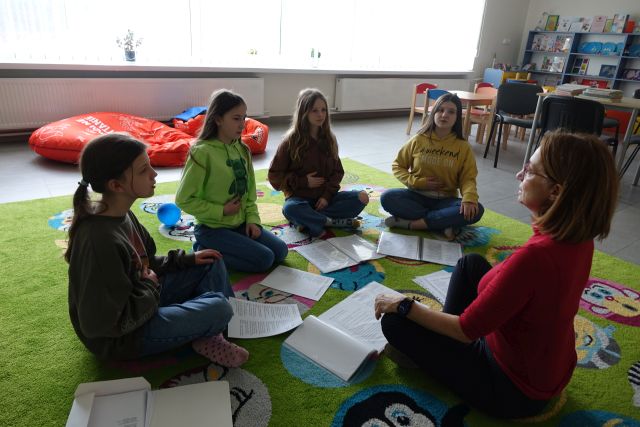 4 dziewczyny i kobieta siedzą na kolorowym dywanie, przed nimi leżą zapisane kartki, trzymają dłonie na brzuchach