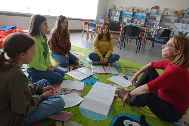 4 dziewczyny i kobieta siedzą na kolorowym dywanie, przed nimi leżą zapisane kartki