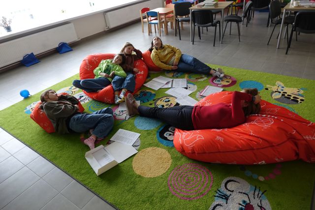 4 dziewczyny i kobieta leżą na pomarańczowych pufach i dywanie, pomiędzy nimi leżą zapisane kartki papieru