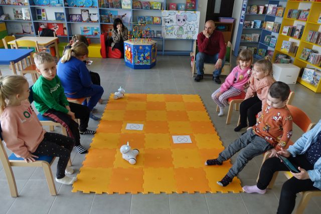 8 dzieci siedzi na krzesełkach, przed nimi na podłodze rołożona pomarańczowa mata, a na niej kartki i 2 roboty Photon