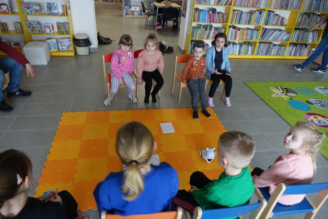 8 dzieci siedzi na krzesełkach, przed nimi na podłodze rołożona pomarańczowa mata, a na niej kartki i 2 roboty Photon