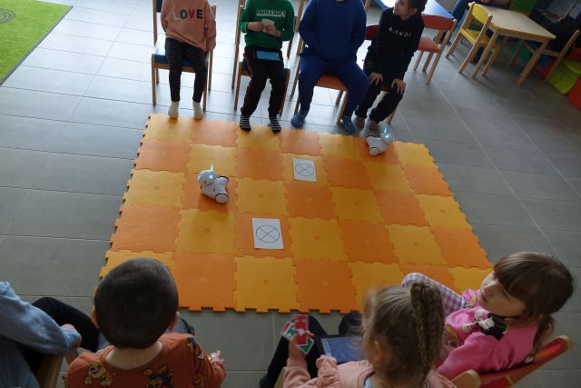 8 dzieci siedzi na krzesełkach, przed nimi na podłodze rołożona pomarańczowa mata, a na niej kartki i 2 roboty Photon