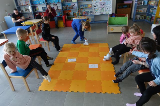 8 dzieci siedzi na krzesełkach, przed nimi na podłodze rołożona pomarańczowa mata, a na niej kartki i 2 roboty Photon